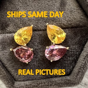 Könnte beinhalten: Ein Paar tropfenförmige Ohrringe mit einem Design aus gelben und violetten Edelsteinen. Die Ohrringe werden in einer grauen Samtbox präsentiert. Der Text "SHIPS SAME DAY" und "REAL PICTURES" ist in Gelb auf der Box abgebildet.