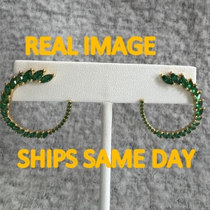 Könnte beinhalten: Ein Paar goldfarbene Creolen, jeweils verziert mit einer Reihe smaragdgrüner, blattförmiger Edelsteine. Die Ohrringe werden auf einem weißen Ständer vor einem strukturierten grauen Hintergrund präsentiert. Der Text "REAL IMAGE" und "SHIPS SAME DAY" sind sichtbar.