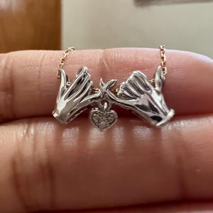 Può includere: Collana color argento con un design a promessa di mignolo, con due mani e un ciondolo a forma di cuore. Il cuore è impreziosito da piccole pietre scintillanti. La catena è di un delicato colore oro.