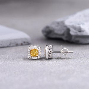 May include: A pair of gold earrings with a square-shaped yellow diamond in the center of each earring. The diamonds are surrounded by a halo of smaller diamonds.