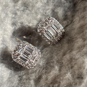 May include: A pair of square-shaped stud earrings. Each earring features a central cluster of rectangular-cut stones surrounded by smaller, round stones. The earrings are set in a silver-colored metal and sparkle brightly.