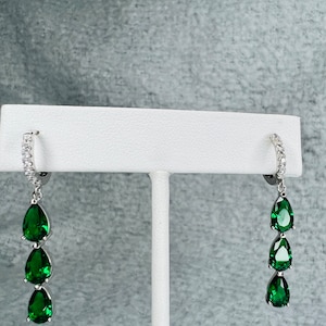 May include: A pair of silver dangle earrings with three emerald green pear-shaped stones on each earring.
