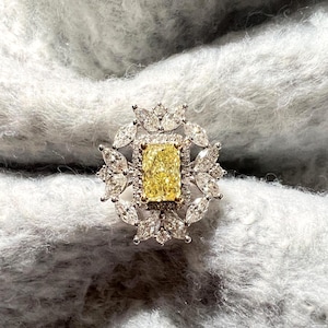 May include: A ring featuring a central rectangular radiant-cut yellow stone set in gold, surrounded by a halo of small round stones and an outer floral-inspired arrangement of marquise-cut white stones set in a polished silver-toned metal band.