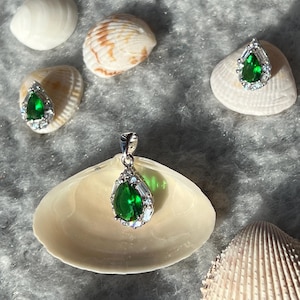 May include: A silver pendant necklace with a teardrop-shaped green gemstone surrounded by smaller clear gemstones. The pendant is displayed on a seashell.