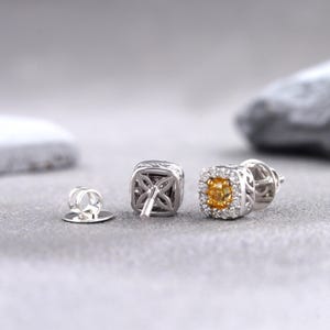 May include: A pair of silver stud earrings with a square setting. The earrings feature a small, round, yellow gemstone surrounded by smaller, clear gemstones. The back of the earrings are visible, showing the post and butterfly clasp.