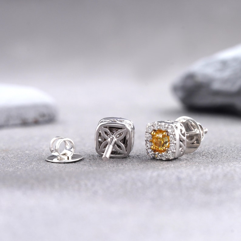 May include: A pair of silver stud earrings with a square setting. The earrings feature a small, round, yellow gemstone surrounded by smaller, clear gemstones. The back of the earrings are visible, showing the post and butterfly clasp.