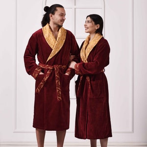 Op de afbeelding: Twee mensen dragen bijpassende bordeauxrode badjassen met gouden bies en gouden gepatroneerde ceinturen. De badjassen hebben een shawlkraag en zijn in de taille gebonden. De mensen staan voor een witte muur met een bloemenkrans die erboven hangt.