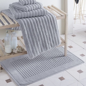 Puede incluir: Un juego de toallas de baño grises y una alfombra de baño a juego con textura acanalada. Las toallas están apiladas en un estante de madera y la alfombra está en el suelo.