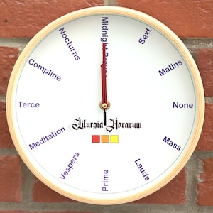 May include: A white circular clock with a wooden frame and Roman numeral markings. The clock face features the text "Liturgia Horarum" and the words "Midnight", "Nocturns", "Compline", "Terce", "Meditation", "Vespers", "Prime", "Lauds", "Sext", "Matins", "None", and "Mass" in a purple font.