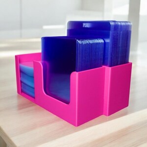 Op de afbeelding: Een felroze bureau-organizer met stapels blauwe plastic vellen. De organizer heeft drie compartimenten, met de vellen netjes gerangschikt. Het woord "PIU" is zichtbaar aan de bovenkant.