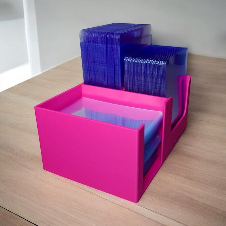 Op de afbeelding: Een felroze bureau-organizer met meerdere compartimenten. Het bevat stapels blauwe kaarten of papieren. Het ontwerp is modern en geometrisch, met een strakke, minimalistische uitstraling. De organizer staat op een licht houten oppervlak.