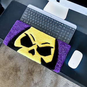 May include: A rectangular rug featuring a yellow skull design with black details and a purple border. The rug is placed on a black surface, next to a keyboard and a white mouse. The rug's design is reminiscent of a popular cartoon character.