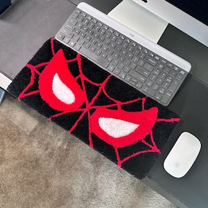 May include: A rectangular black rug with a red and white spider-eye design. The rug is placed on a desk next to a keyboard and a white mouse. The rug's design features a red spider-web pattern.