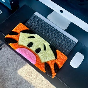 May include: A rectangular, handmade rug featuring a cartoon face design. The rug has an orange border with black accents, a light green center, a pink curved shape, and black oval shapes. A keyboard and mouse are next to the rug.