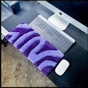 Op de afbeelding: Een paars-blauwe gestreepte polsensteun voor toetsenbord met een grijs toetsenbord en een witte muis op een zwarte bureaumat.