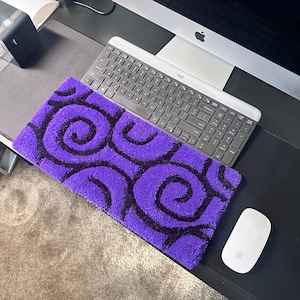 May include: A rectangular purple rug with a black spiral pattern. The rug is placed on a desk next to a computer keyboard and mouse. The rug's dimensions are approximately 24 inches long and 12 inches wide.