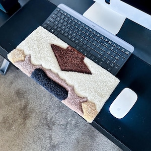 May include: A rectangular, textured rug with a mountain-like design. The rug features a cream-colored base with brown, navy, pink, and tan accents. A keyboard and mouse are visible in the background, suggesting a desk setting.