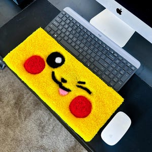 Op de afbeelding: Een gele, pluche polsensteun voor het toetsenbord met een cartoon gezicht van Pikachu, een populair Pokémon-personage. Het gezicht heeft zwarte ogen, een rode neus en een roze tong die uitsteekt. De polsensteun ligt op een zwart bureau met een toetsenbord en een muis.