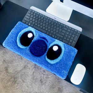 Op de afbeelding: Een blauw tapijt met het gezicht van een stripfiguur met grote zwarte ogen, witte highlights en een donkerblauwe neus. Een draadloos toetsenbord en muis liggen op een zwart bureau.