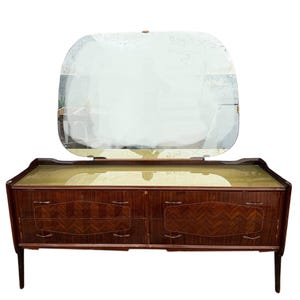 Puede incluir: Un tocador de madera vintage con un gran espejo ovalado. El tocador tiene cuatro cajones con un patrón de veta de madera y una parte superior de color verde claro. El tocador está sobre cuatro patas.