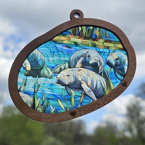 May include: Wooden ornament featuring a stained-glass style depiction of manatees in a blue and green aquatic scene. The ornament has a brown wooden frame and the text "Floaty Potato".