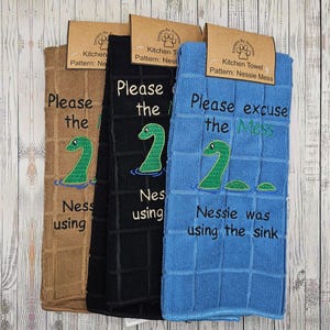 May include: Three kitchen towels with a humorous embroidered design featuring a green Nessie monster. The brown towel reads "Please the Mess Nessie was using". The black towel reads "Please the Mess Nessie was using". The blue towel reads "Please excuse the Mess Nessie was using the sink".