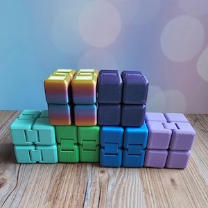 May include: A collection of colorful fidget cubes in shades of teal, green, blue, purple, and a rainbow gradient. The cubes are arranged in a stacked formation on a wooden surface, with a soft blue and purple background.