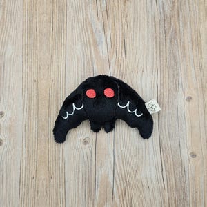 May include: A black plush moth toy with red eyes and white wing details. The moth has a small tag attached. The toy is made of soft material and is designed to resemble a moth.