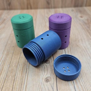 May include: Three cylindrical containers in green, purple, and blue, with a textured surface. The blue container is disassembled, showing the lid and body. Each container has a series of small holes and a button-like design on the lid.