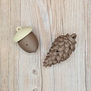 May include: Two brown decorative items: an acorn and a pinecone. The acorn has a light tan cap and a dark brown body. The pinecone is a darker brown with a textured surface. Both items have a small hole.