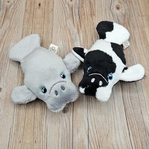 May include: Two plush toys: a gray manatee and a black and white cow. Both have embroidered blue eyes and are lying on a wooden surface. The manatee is light gray, and the cow has black spots.