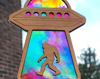 May include: A wooden UFO ornament with a rainbow-colored stained glass design. The UFO is hovering over a silhouette of Bigfoot.