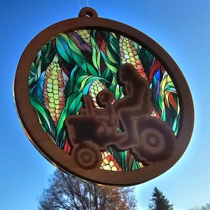 May include: A round wooden ornament with a stained glass design of corn stalks and a tractor silhouette. The ornament is brown with a clear glass center.