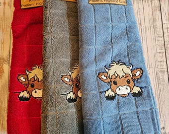 Puede incluir: Tres toallas de cocina con un diseño de vaca Highland. Las toallas son rojas, grises y azules. Cada toalla tiene el texto "Kitchen Towel" y "Pattern: Highland Cow" impreso en la etiqueta.