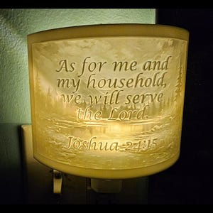 May include: A cream-colored night light with the text "As for me and my household, we will serve the Lord. Joshua 24:15." The night light features a landscape scene and is plugged into a wall outlet.