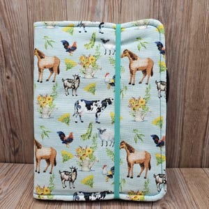 May include: A light blue fabric journal cover featuring a farm animal pattern. The cover has images of horses, cows, goats, roosters, and sheep. Yellow sunflowers in white vases are also present. A teal elastic band secures the cover.