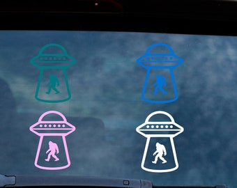 May include: Four vinyl stickers featuring a UFO design with a Bigfoot silhouette inside. The stickers are in teal, blue, pink, and white. The stickers are designed to be applied to a car window.