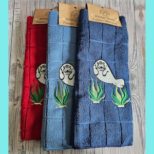 May include: Three kitchen towels with a manatee design. The towels are red, blue, and light blue. The manatee is embroidered in white with green seaweed. The towels have the text "Kitchen Towel Pattern: Manatee" on the tag.