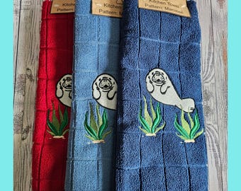 Puede incluir: Tres toallas de cocina con un diseño de manatí. Las toallas son rojas, azules y azul claro. El manatí está bordado en blanco con algas verdes. Las toallas tienen el texto "Kitchen Towel Pattern: Manatee" en la etiqueta.