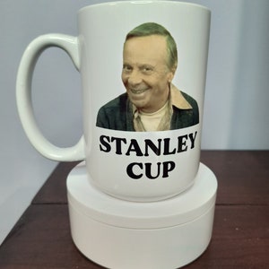 May include: White ceramic mug featuring a picture of a man and the text "Stanley Cup" in black lettering.