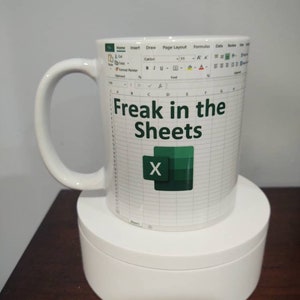 May include: White ceramic mug with a green and white Microsoft Excel spreadsheet design. The mug has a handle and the text "Freak in the Sheets" and the letter "X" are printed on the front.