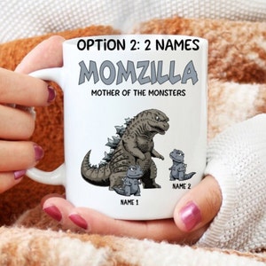 May include: White ceramic coffee mug with a Godzilla-themed design. The mug reads "Momzilla, Mother of the Monsters" with a cartoon image of Godzilla and two smaller Godzilla figures. The text "Name 1" and "Name 2" are at the bottom of the mug.