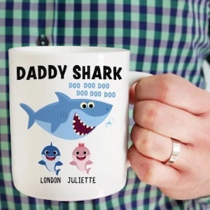May include: White ceramic mug with a blue shark illustration and the text "Daddy Shark Doo Doo Doo Doo Doo Doo Doo" and the names "London Juliette".