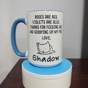 May include: White ceramic mug with a blue rim and handle. The mug has a black illustration of a cat with the text "Roses are red, violets are blue. Thanks for feeding me and scooping up my poo. Love, Shadow."