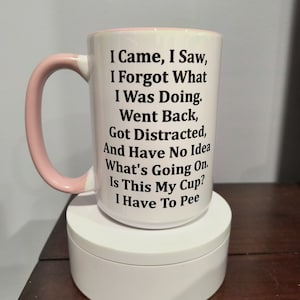 Può includere: Una tazza in ceramica bianca con un manico rosa. La tazza ha un testo nero che recita "Sono venuto, ho visto, ho dimenticato cosa stavo facendo. Sono tornato indietro, sono stato distratto e non ho idea di cosa stia succedendo. È questa la mia tazza? Devo fare pipì."
