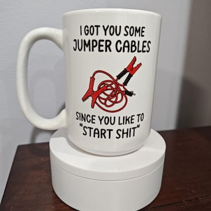 May include: White ceramic coffee mug with a black and red illustration of jumper cables. The text on the mug reads "I got you some jumper cables since you like to "start shit""