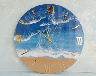 May include: Circular wall clock depicting a beach scene. The clock face shows a blue ocean with white waves and a sandy shore. Gold-coloured Roman numerals and clock hands complete the design.