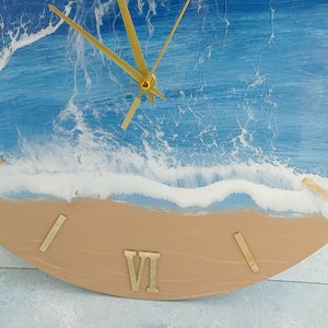 Puede incluir: Un reloj de pared redondo con un dise&ntilde;o de escena de playa. La esfera del reloj presenta un oc&eacute;ano azul con olas blancas y una playa de arena. Las manecillas y los n&uacute;meros romanos dorados completan el dise&ntilde;o.
