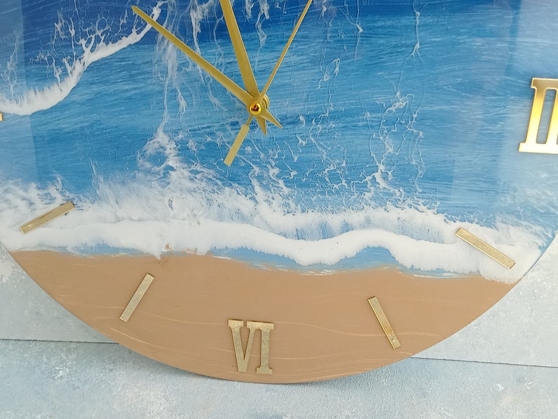 Puede incluir: Un reloj de pared redondo con un dise&ntilde;o de escena de playa. La esfera del reloj presenta un oc&eacute;ano azul con olas blancas y una playa de arena. Las manecillas y los n&uacute;meros romanos dorados completan el dise&ntilde;o.