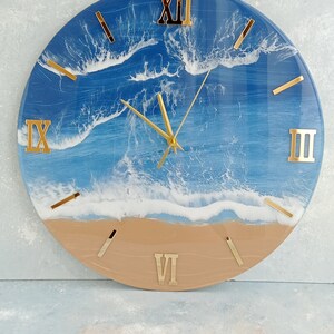Puede incluir: Reloj de pared redondo con tem&aacute;tica de playa. La esfera del reloj presenta un dise&ntilde;o de oc&eacute;ano azul con olas blancas y una playa beige en la parte inferior. Los n&uacute;meros romanos y las manecillas doradas completan el dise&ntilde;o.